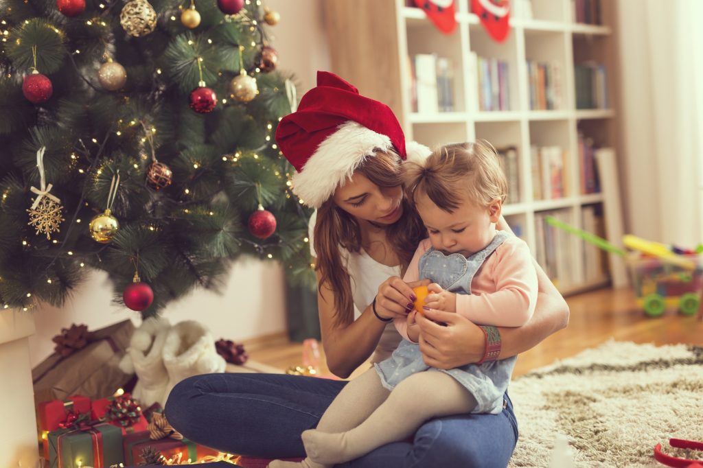 Young mother holding her baby girl in the lap, sitting on the floor next to a nicely decorated Christmas tree and peeling a tangerine