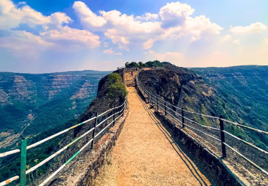 A narrow concrete pathway with white railings extending towards a cliff edge, overlooking a vast expanse of blue water or sky with white clouds