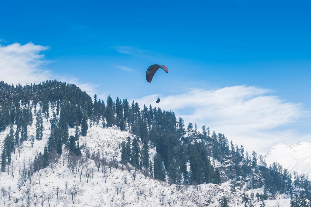 Tandem paraglider soaring over snow-covered Solang Valley near Manali in Himachal Pradesh against a clear blue sky