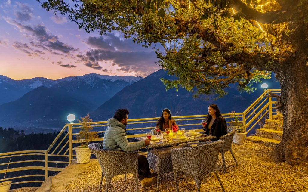 Guests enjoying an outdoor dinner under a tree at a SaffronStays Interstellar in Manali with snow-capped Himalayan peaks at sunset.