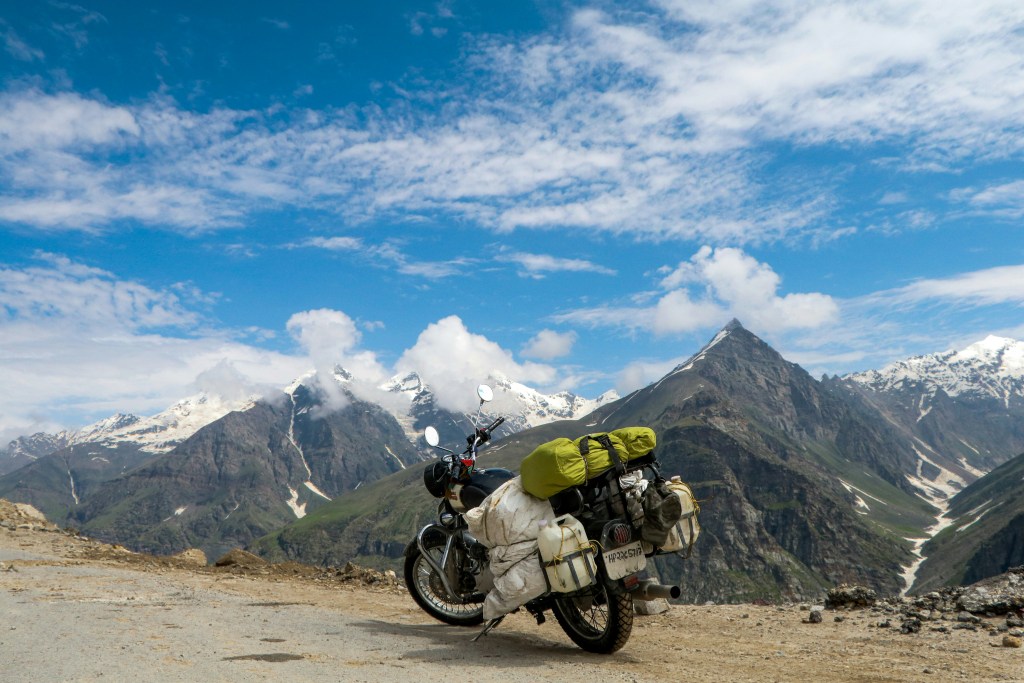 Loaded Royal Enfield motorcycle parked at Rohtang Pass near Manali with snow-capped Himalayan peaks and blue sky in the background