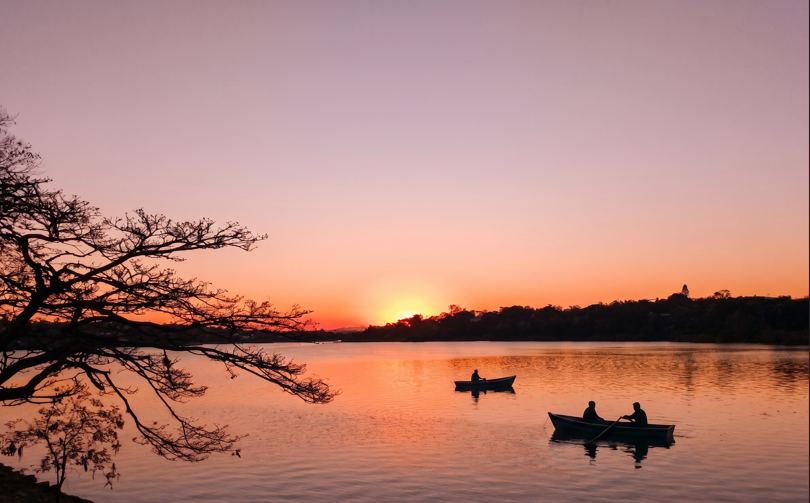 Wide shot of Pawna Lake at golden hour or under monsoon cloud cover, with Tikona or Lohagad fort visible in the background, tents or campsite silhouettes optional in the foreground.