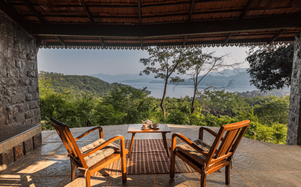 Mulshi backwaters shot from one of SaffronStays Sunkissed, grey monsoon sky, still water reflecting the hills, a couple or small group seated at an outdoor table optional for scale.