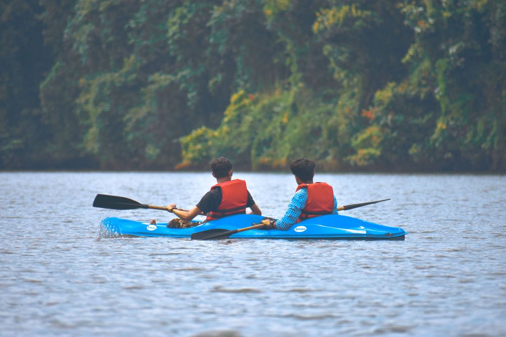 Panshet Dam backwaters at high water, water sports activity (kayaking or boating) visible in the middle distance, green hills surrounding the reservoir, overcast sky.