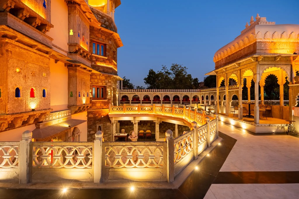  SaffronStays The Shikargarh Palace heritage exterior with original stone Odi structure at centre, gardens and pool visible, Aravalli hills backdrop.