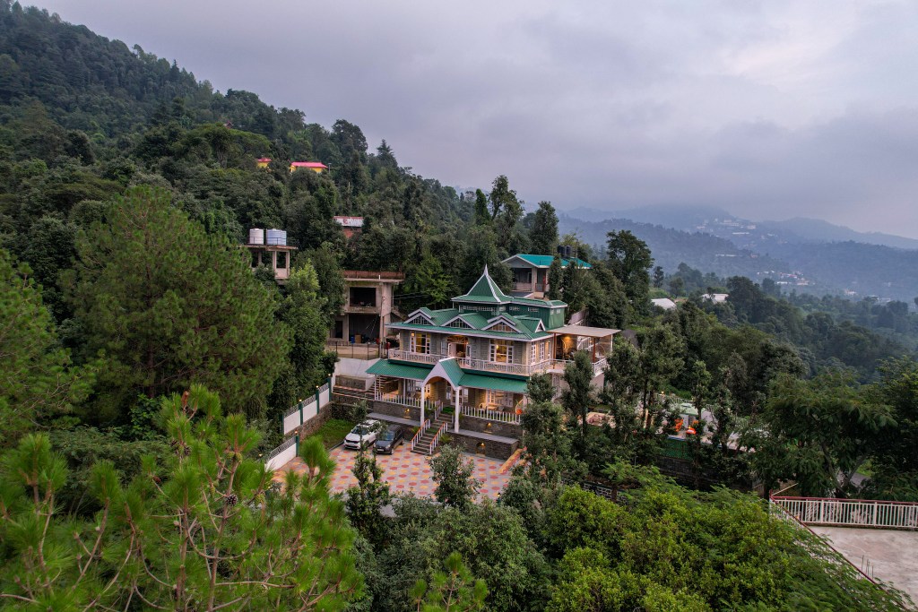 Aerial view of a modern house surrounded by dense greenery and mountains, with a driveway and parking area.