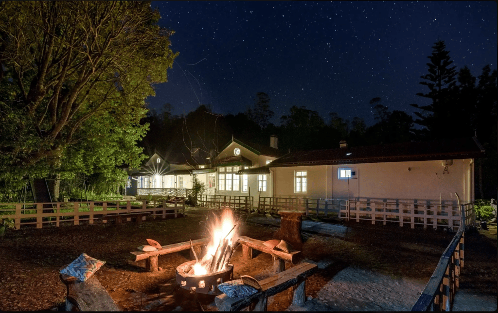 A night view of a rustic building surrounded by trees, featuring a campfire at the forefront and a starry sky overhead.