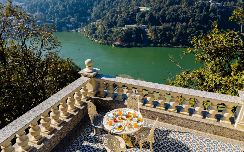 Scenic view from a terrace overlooking a green lake surrounded by hills, featuring a beautifully set table with a variety of breakfast items.