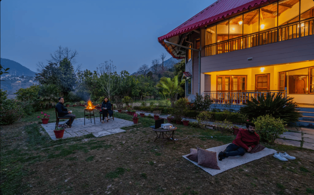 Two people sitting around a fire pit in a garden, with a house in the background, during twilight.