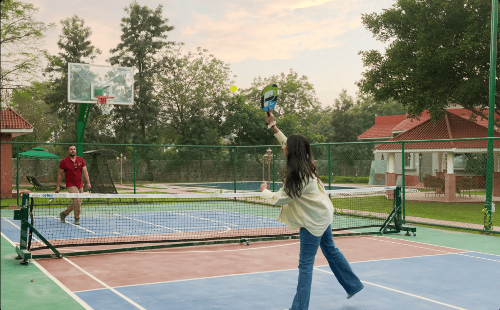 A young girl holding a paddle preparing to serve in a game on a multi-sport court, with a basketball hoop visible in the background.