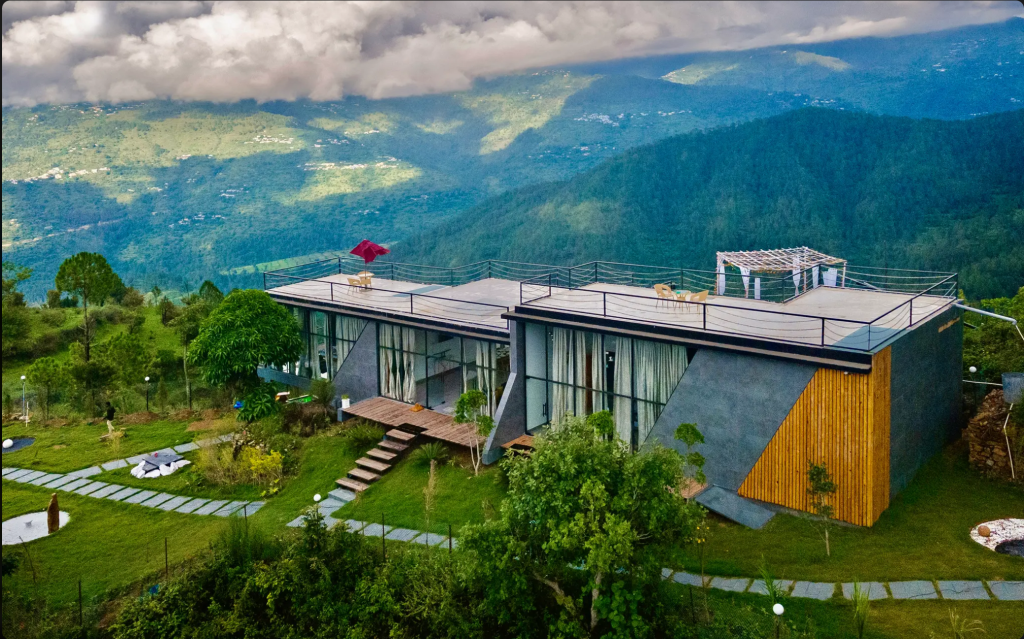 A modern house with large glass windows and a flat roof, situated on a hillside with lush greenery and mountains in the background.
