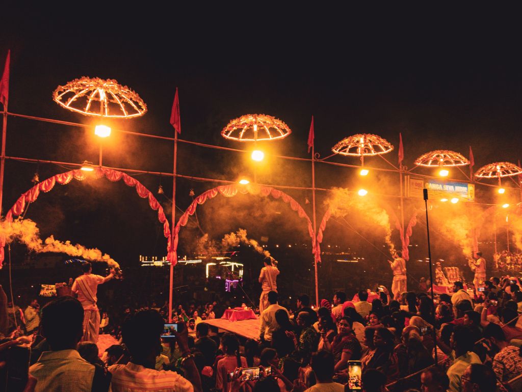 A lively night scene from a cultural festival, featuring performers in traditional attire, smoke billowing, and illuminated decorations with hanging lights and banners, surrounded by a crowd capturing the moment with smartphones.