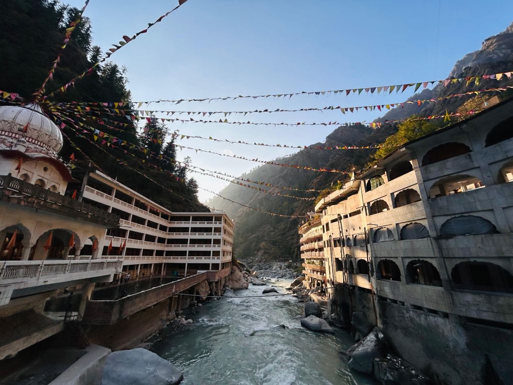 View of a river flowing between buildings under a clear blue sky, with colorful prayer flags strung overhead, surrounded by mountains.
