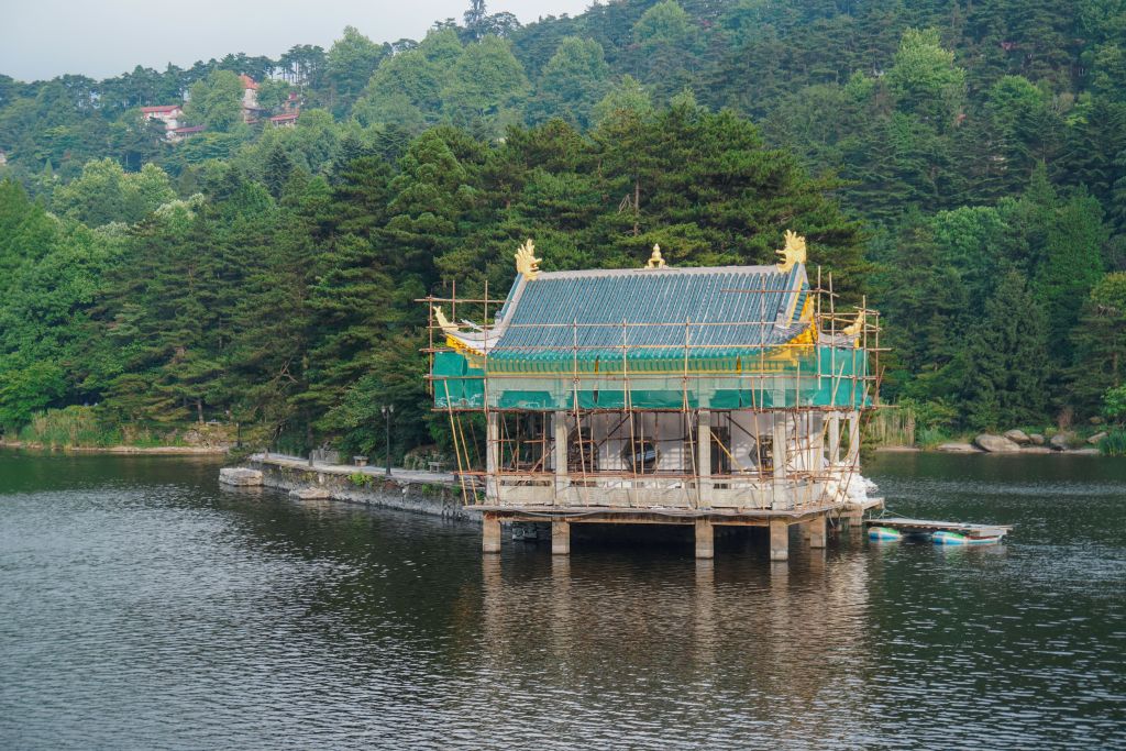 A temple under construction on stilts in the middle of a lake, surrounded by lush green trees and hills.
