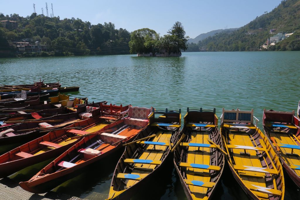 Colorful boats lined up along the shore of a tranquil lake, surrounded by green hills and trees.
