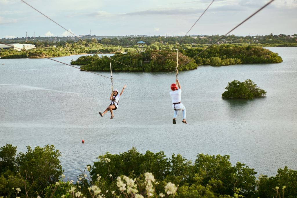 Two individuals ziplining over a body of water surrounded by greenery, with one person raising their arm in excitement.