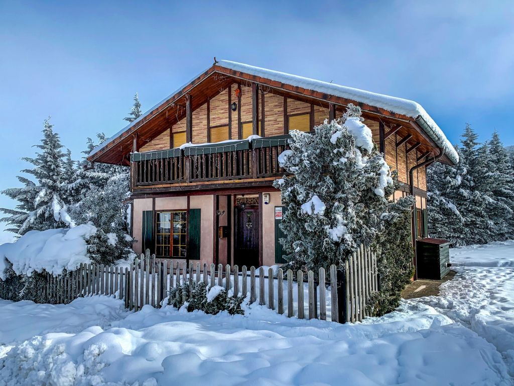 A charming snow-covered cabin surrounded by trees, with a balcony and a fence, under a clear blue sky.