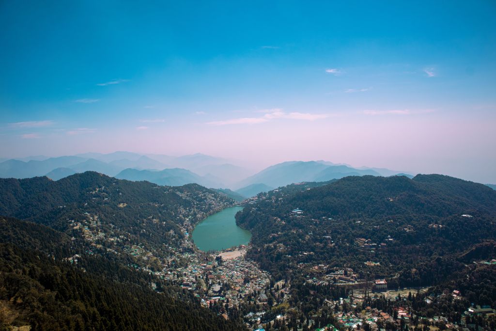 A panoramic view of a lake nestled between mountains, surrounded by a small town and lush greenery under a clear blue sky.