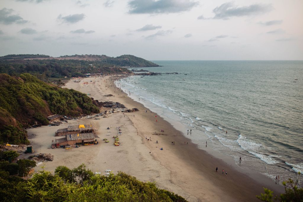 A scenic view of a beach with gentle waves lapping on the shore, surrounded by greenery and hills in the background. People can be seen walking along the beach and enjoying the water.