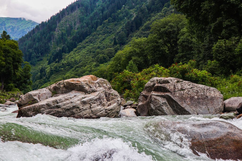 A scenic view of a river flowing over large rocks, surrounded by lush green mountains and trees under a cloudy sky.