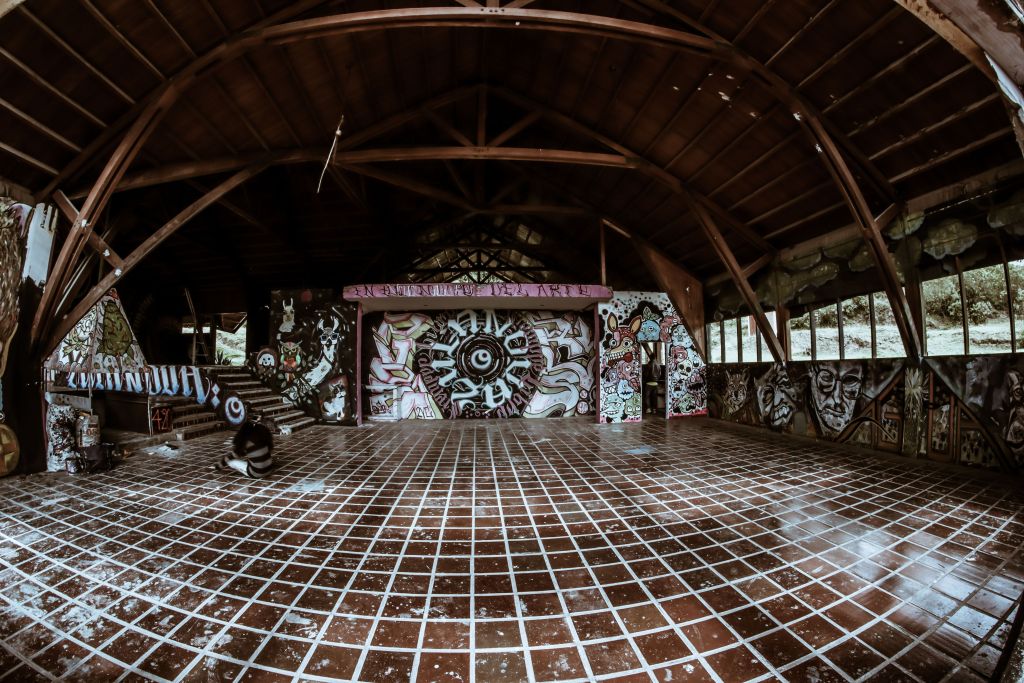 An empty indoor space featuring an expansive tiled floor with a grid pattern, surrounded by colorful graffiti art on the walls and a wooden beam ceiling.