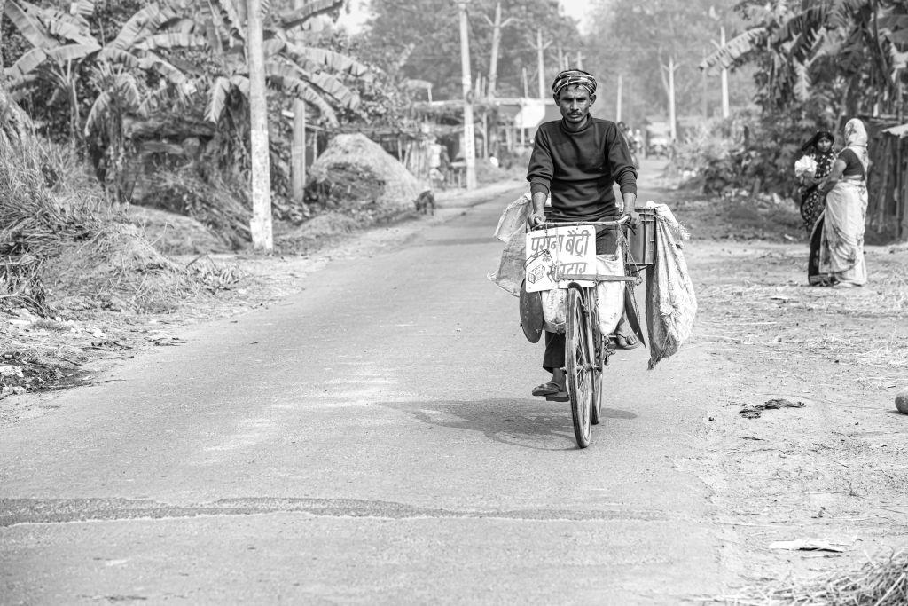 A man riding a bicycle on a rural road, carrying bags and a sign in Hindi, with a few women standing nearby.