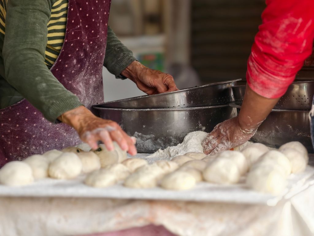 Two hands working with dough on a floured surface, with freshly shaped dough pieces in the foreground and metal containers in the background.