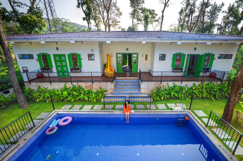 A vibrant outdoor setting featuring a blue swimming pool, surrounded by lush greenery and a well-decorated house with green doors and windows. A woman in an orange outfit sits by the pool, with a staircase leading up to the house.