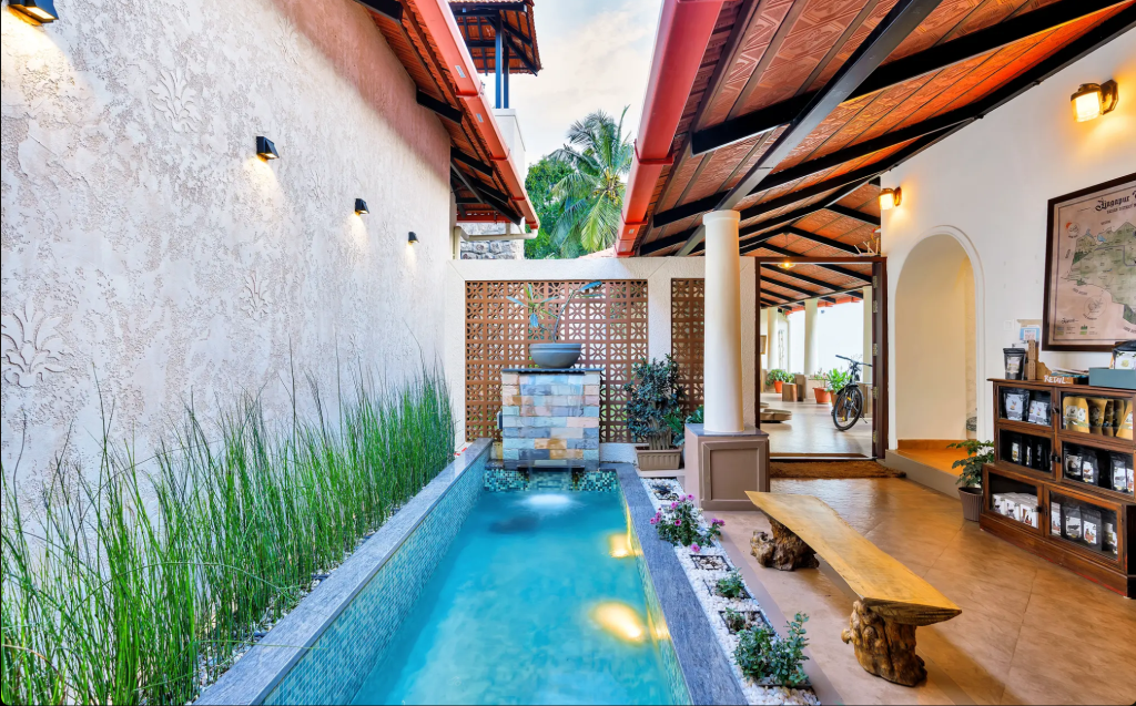 A serene indoor-outdoor space featuring a small pool lined with plants, stone accents, and a wooden bench, with an interior view showing warm lighting and decorative elements.
