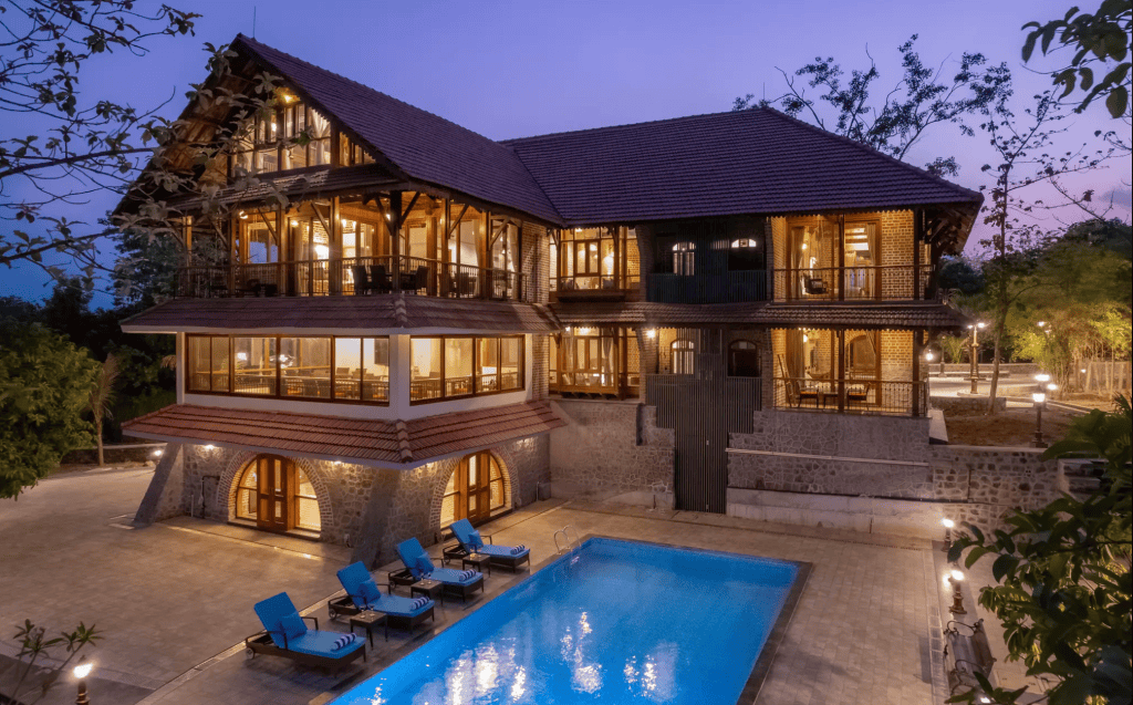 A modern wooden house with large windows and a sloped roof, featuring a swimming pool and lounge chairs in the foreground, set against a twilight sky.