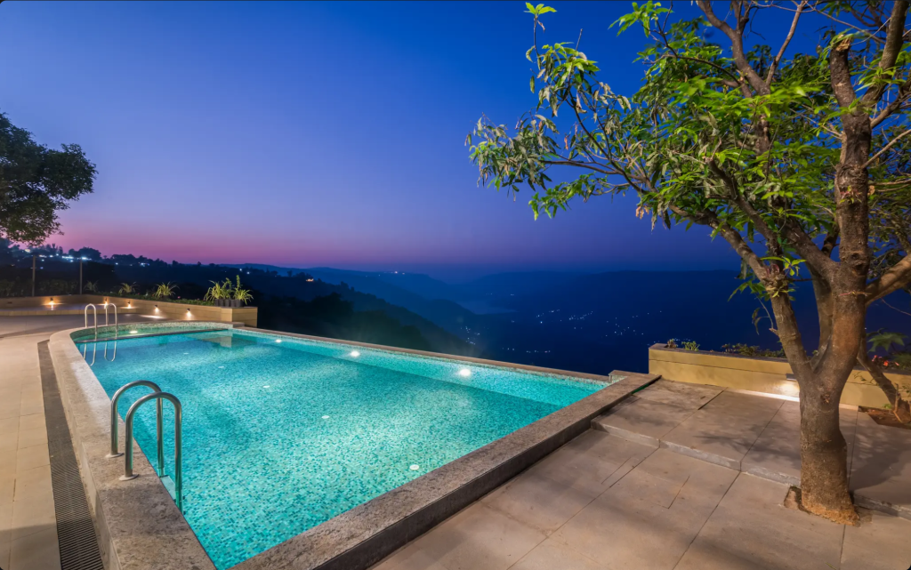 A serene outdoor swimming pool overlooking a mountainous landscape at dusk, illuminated by pool lights and surrounded by lush greenery.