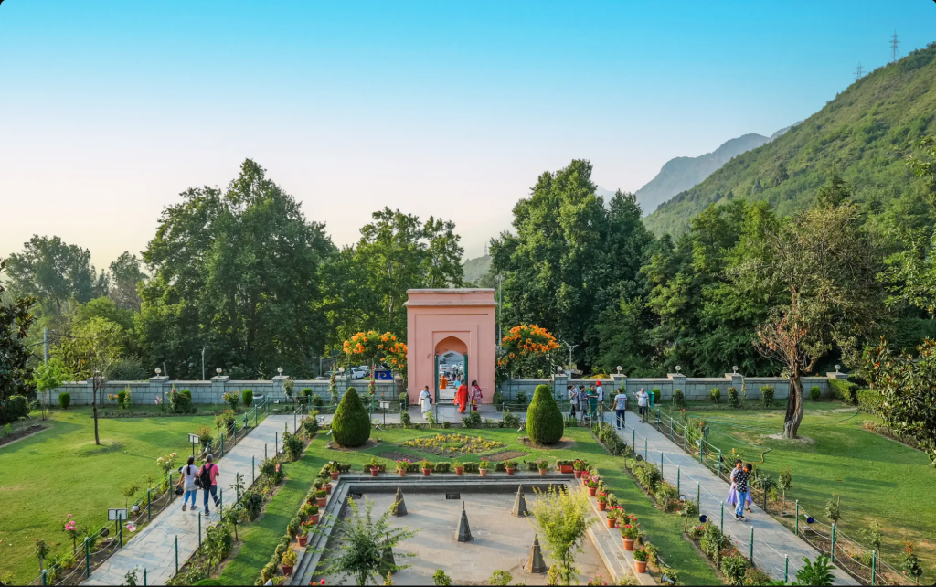 A scenic view of a park featuring well-maintained gardens, pathways, and an ornate structure in the background, surrounded by lush trees and mountains under a clear blue sky.