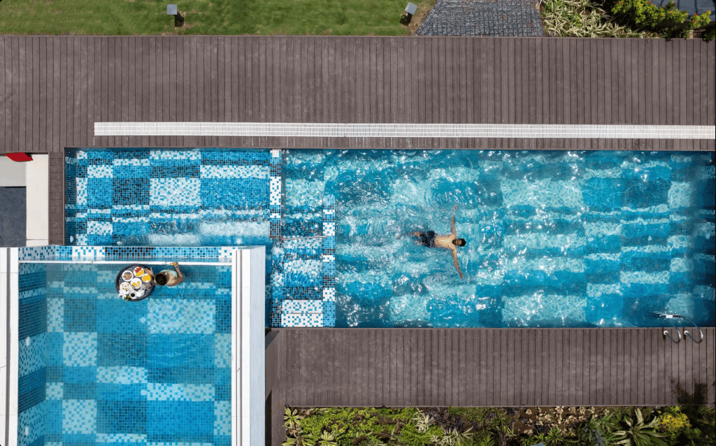 Aerial view of a swimming pool with a person swimming in the water, surrounded by wooden decking and landscaped greenery.