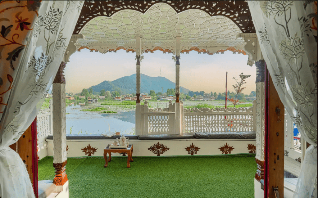 View from a wooden balcony with intricate carvings overlooking a scenic lake and mountains in the background. A table set for tea is visible on a green carpet.