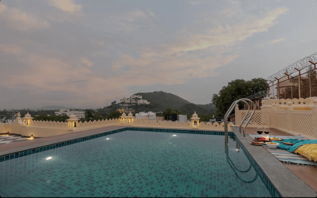 Scenic view of a rooftop swimming pool with turquoise tiles, surrounded by traditional architecture, overlooking a lush landscape and distant hills at dusk.