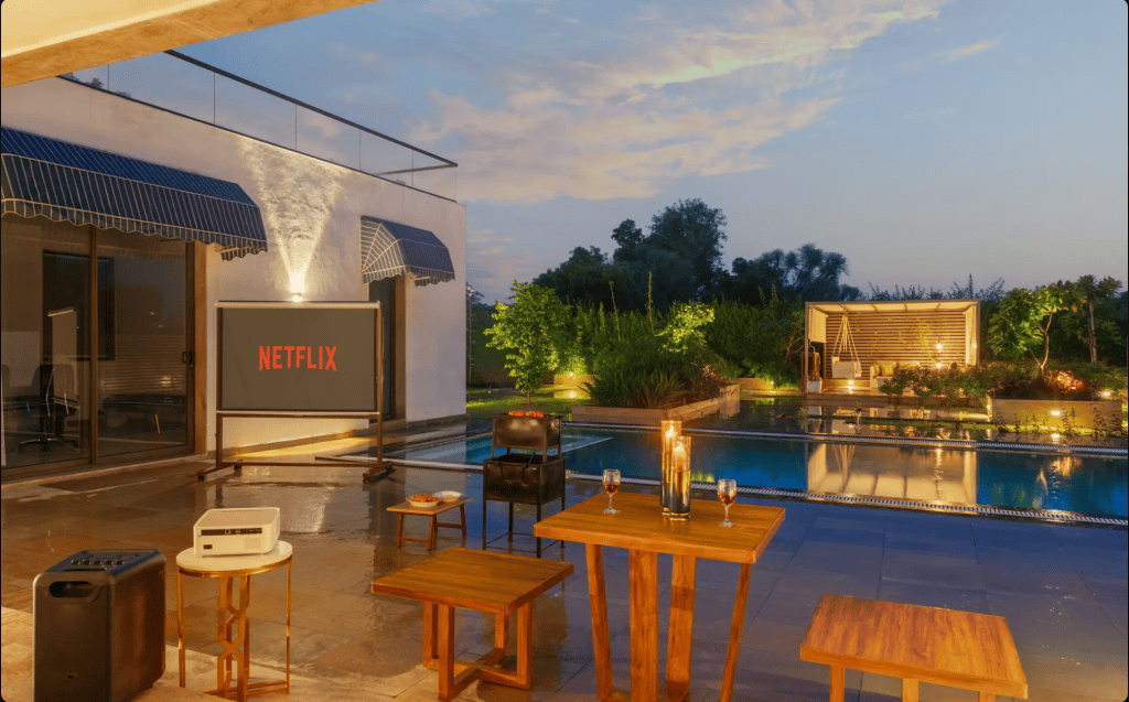 An outdoor movie setup featuring a large screen displaying the Netflix logo beside a swimming pool, with cozy seating arrangements, decorative candles, and lush greenery in the background during twilight.