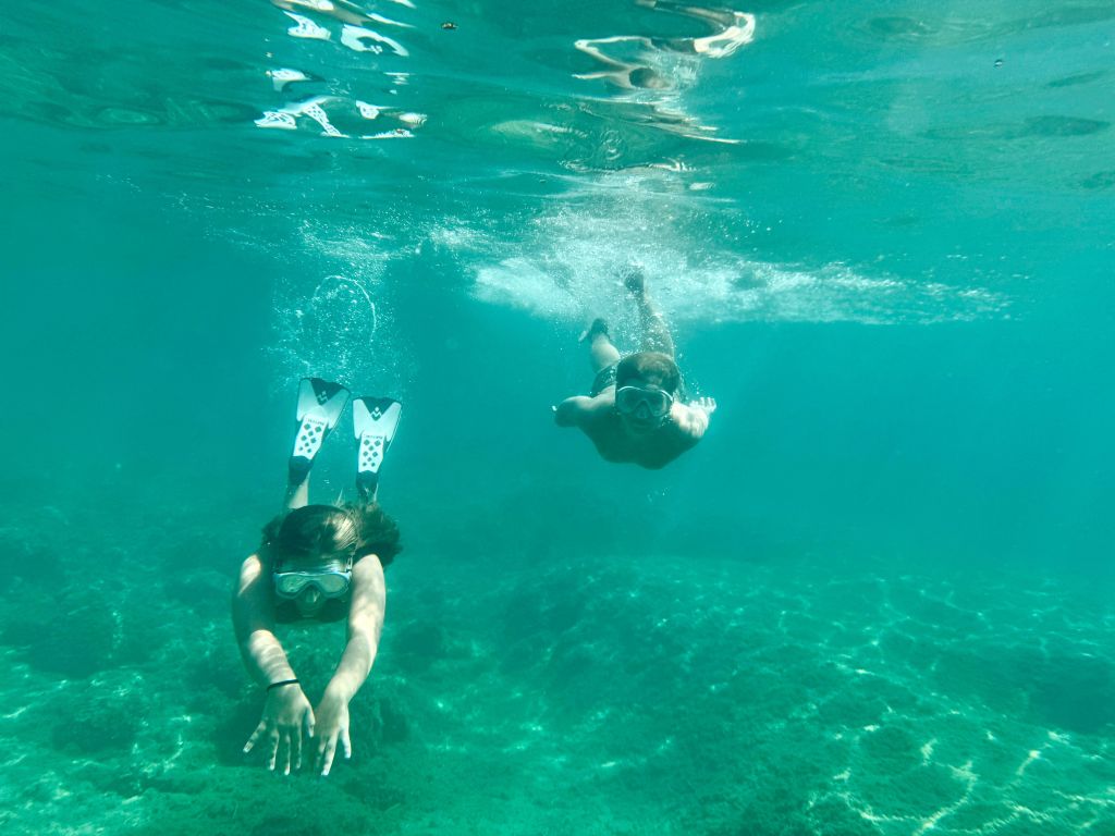 Two individuals swimming underwater in a clear, turquoise sea. One person is wearing snorkel fins and a mask, reaching forward, while the other swims nearby.