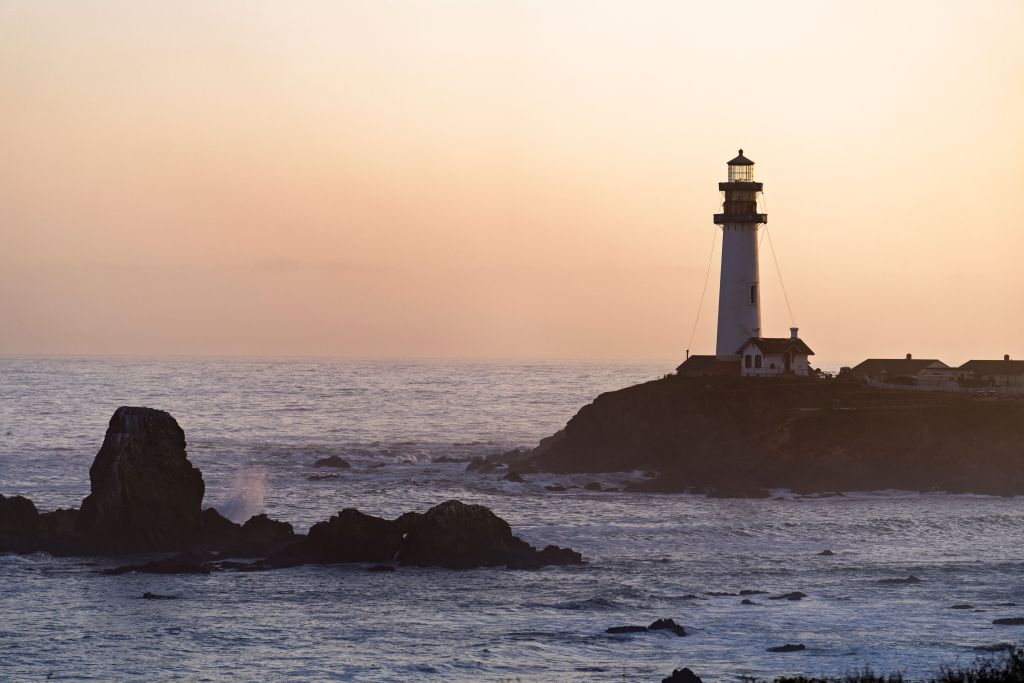 A silhouette of a lighthouse at sunset, standing on a rocky coastline with waves crashing nearby and a soft pastel sky.