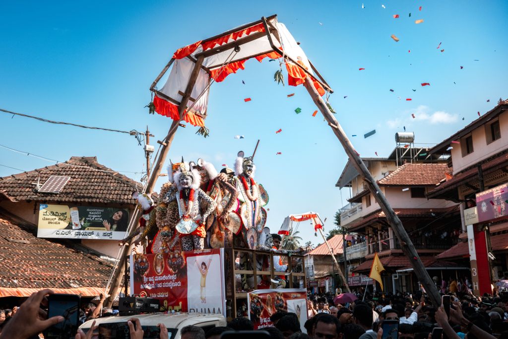 A colorful festival parade featuring performers in traditional costumes on a decorated float, surrounded by a lively crowd and buildings, with confetti falling from the sky.