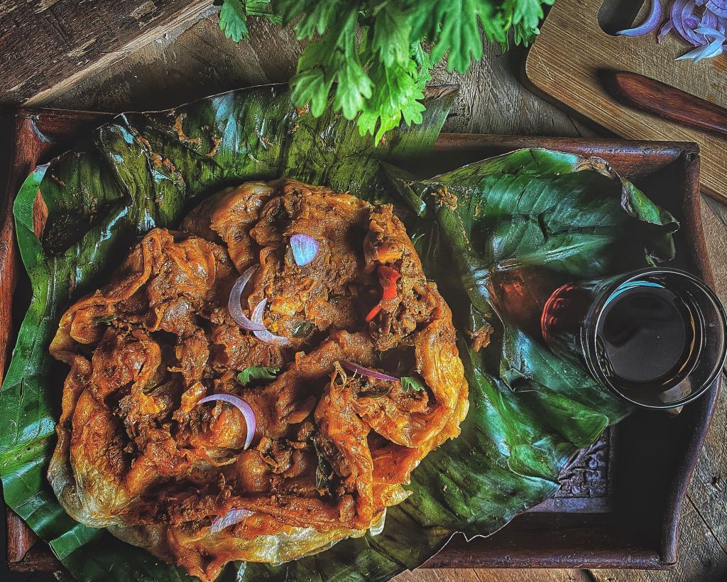 A traditional dish served on banana leaves, featuring marinated meat garnished with onions and herbs, accompanied by a small glass of dark liquid.