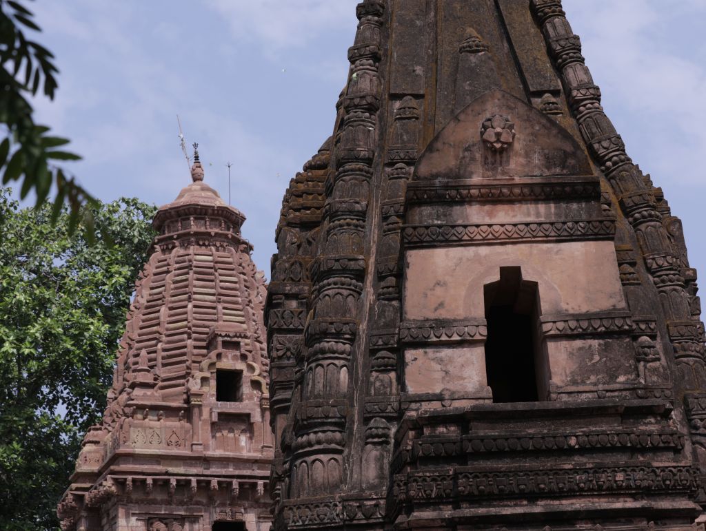 Two intricately designed Hindu temple towers surrounded by greenery under a blue sky.