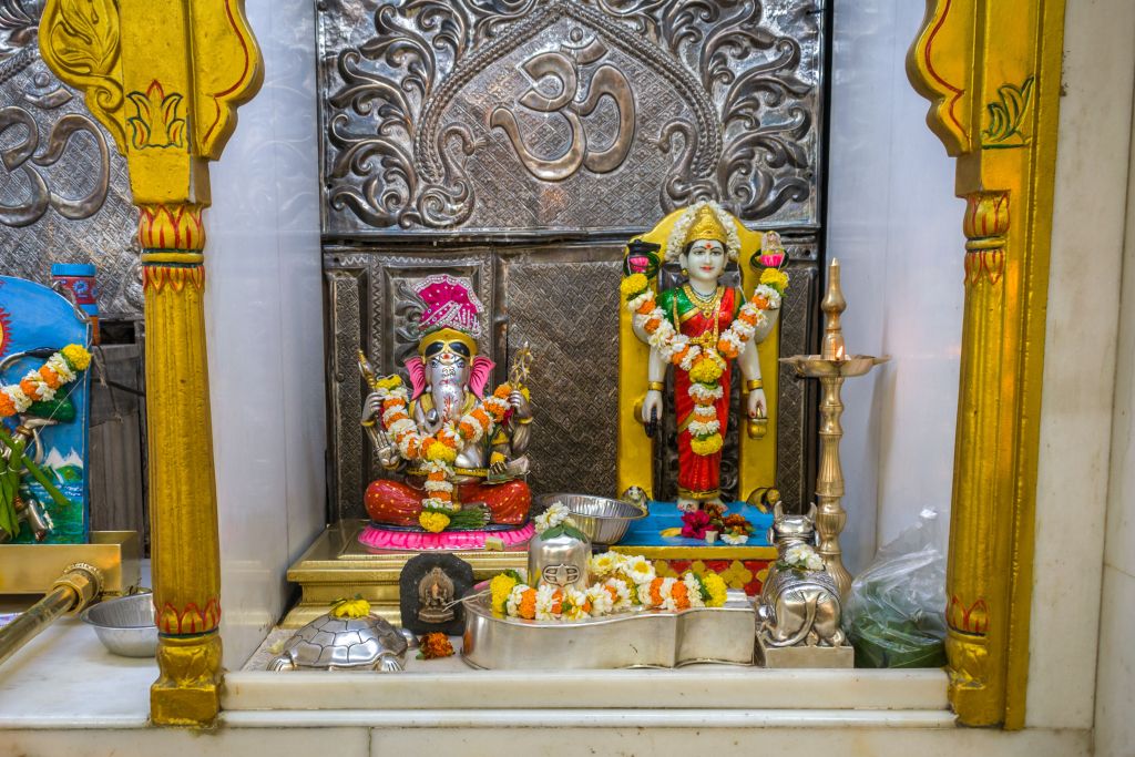 A Hindu shrine featuring statues of deities, including Lord Ganesha and Goddess Lakshmi, adorned with flower garlands and surrounded by decorative elements and worship items.