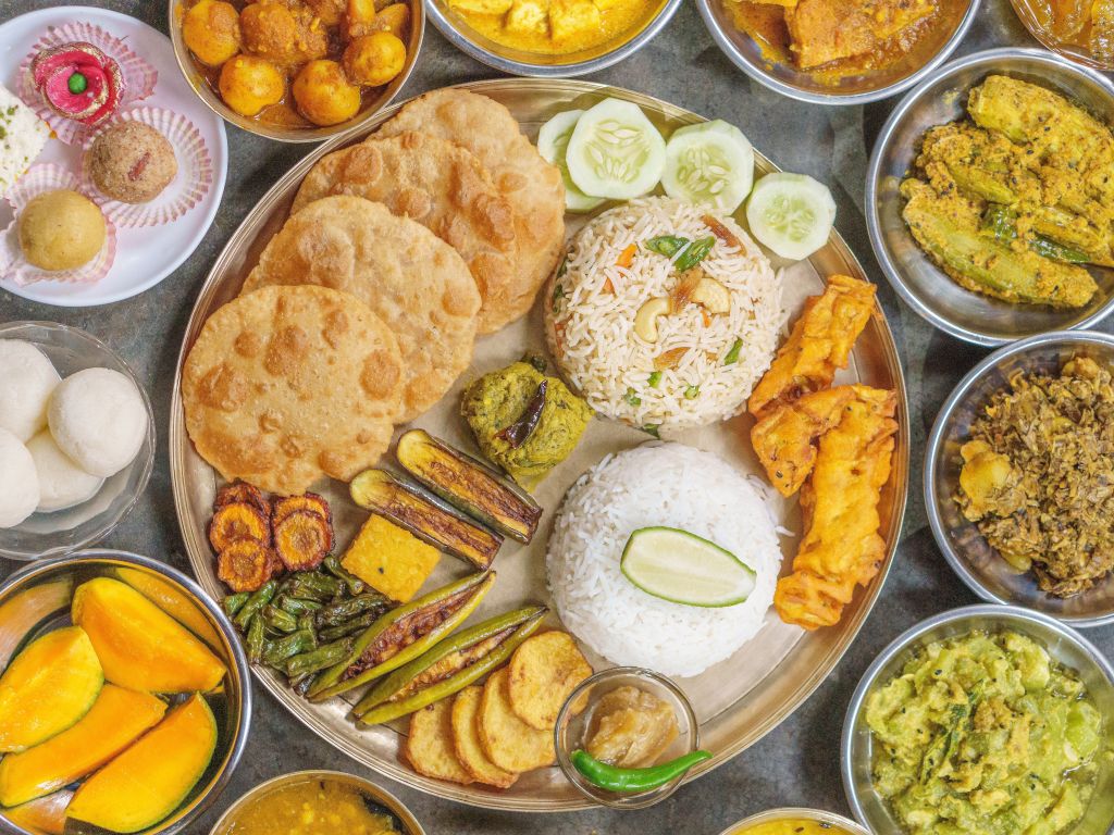 A traditional Indian meal served on a platter, featuring an assortment of dishes including rice, curries, deep-fried snacks, and assorted sweets. The meal includes multiple types of bread, vegetables, and a side of mango slices.