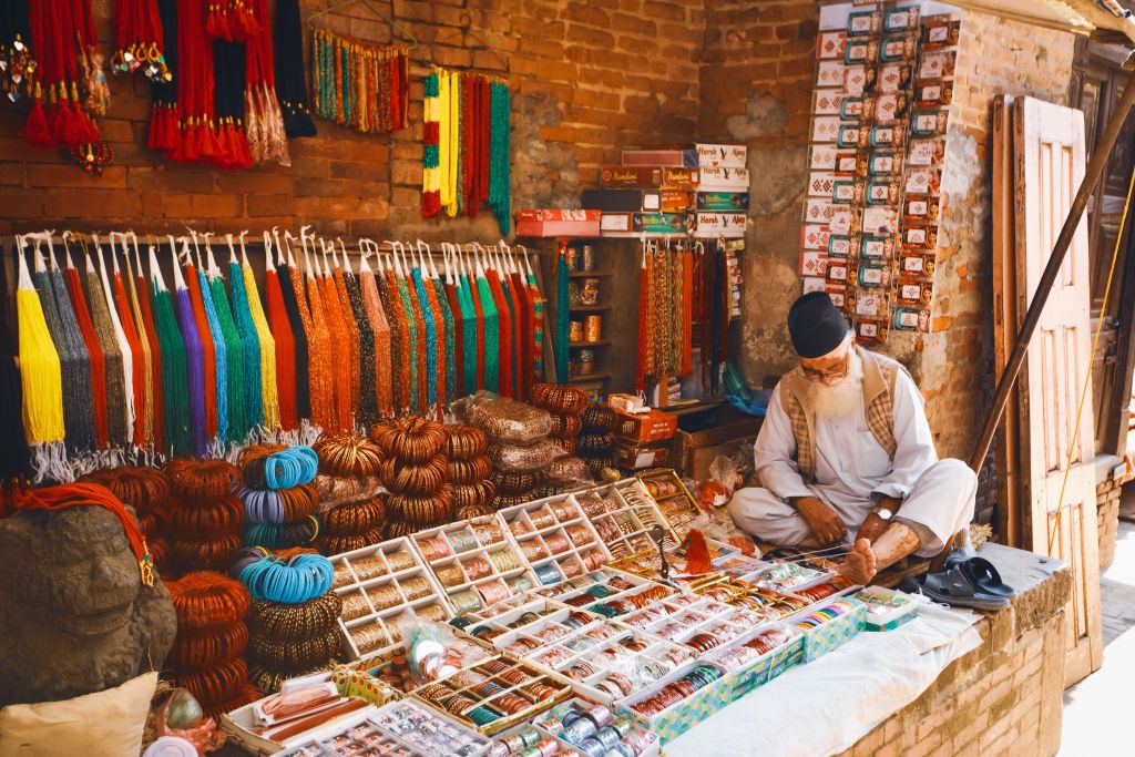A market stall displaying colorful textiles and accessories, with an elderly man sitting on the floor, examining items.