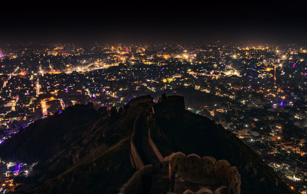 View of a city illuminated at night, taken from a high vantage point with a fort and hills in the foreground.