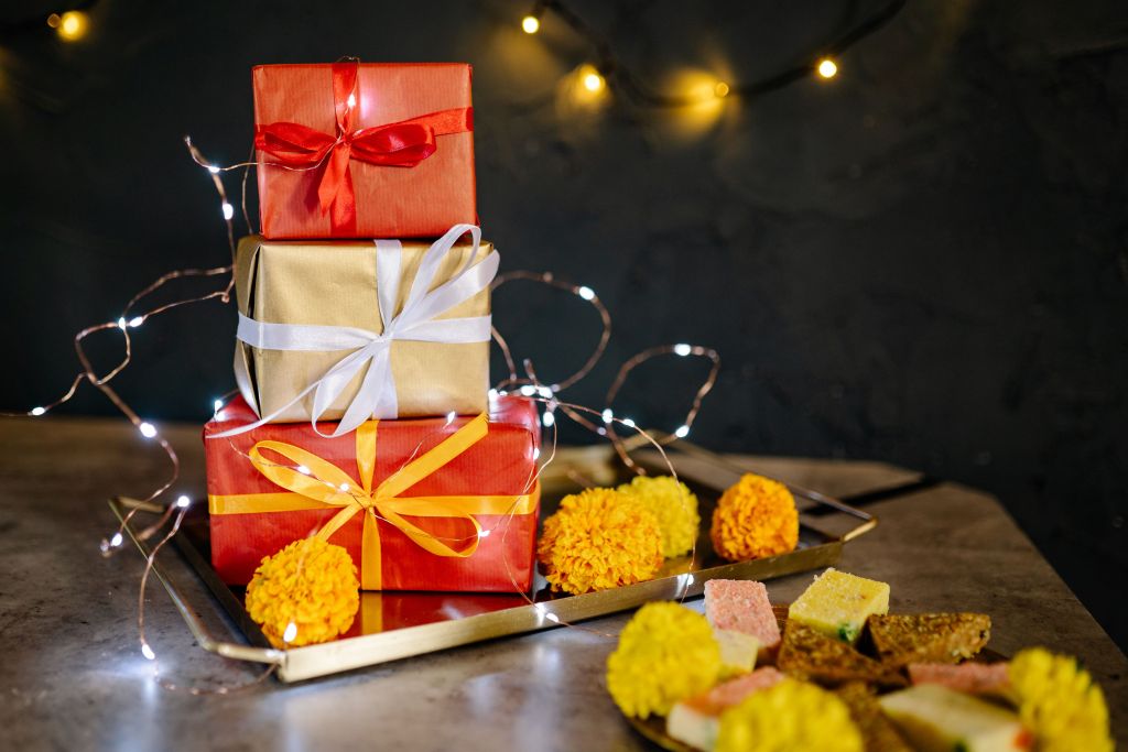 A stack of three decorative gift boxes in red and gold, adorned with colorful ribbons, surrounded by marigold flowers and traditional sweets on a tray, with soft fairy lights in the background.