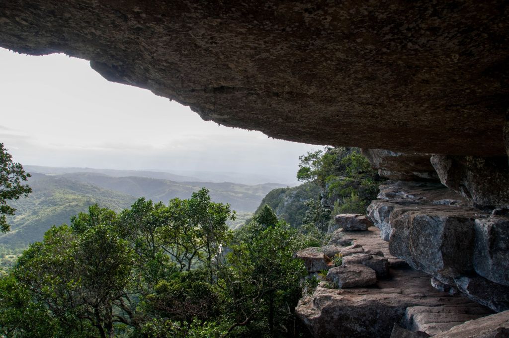 A scenic view from beneath a rocky overhang, showcasing lush green hills and valleys in the background under a cloudy sky.